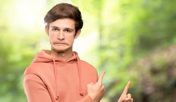 Teenager man with sweatshirt frightened and pointing to the side at outdoors