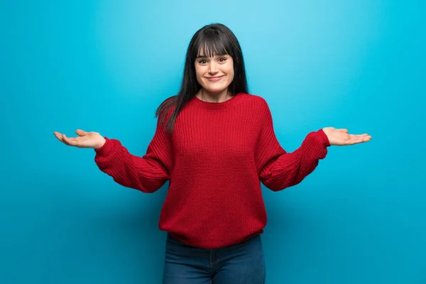 Woman with red sweater over blue wall having doubts while raising hands