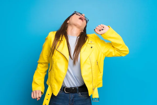 Young woman with yellow jacket on blue background yawning and covering wide open mouth with hand