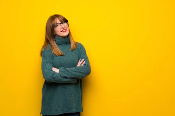 Woman with glasses over yellow wall keeping the arms crossed while smiling