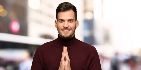 Man with turtleneck sweater keeps palm together. Person asks for something in the city