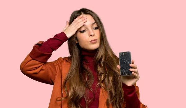 Young woman with coat with troubled holding broken smartphone over isolated pink background