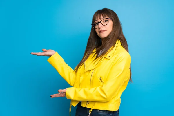Young woman with yellow jacket on blue background extending hands to the side for inviting to come