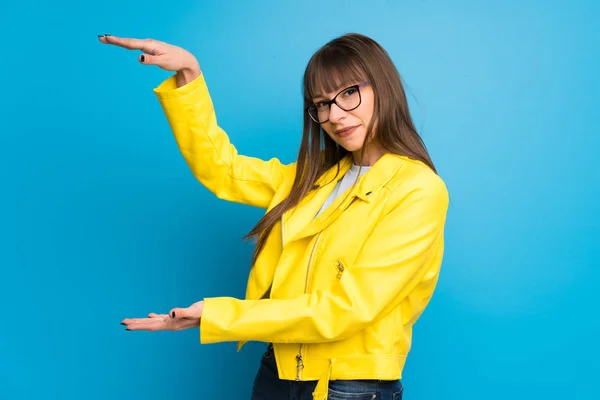 Young woman with yellow jacket on blue background holding copyspace to insert an ad