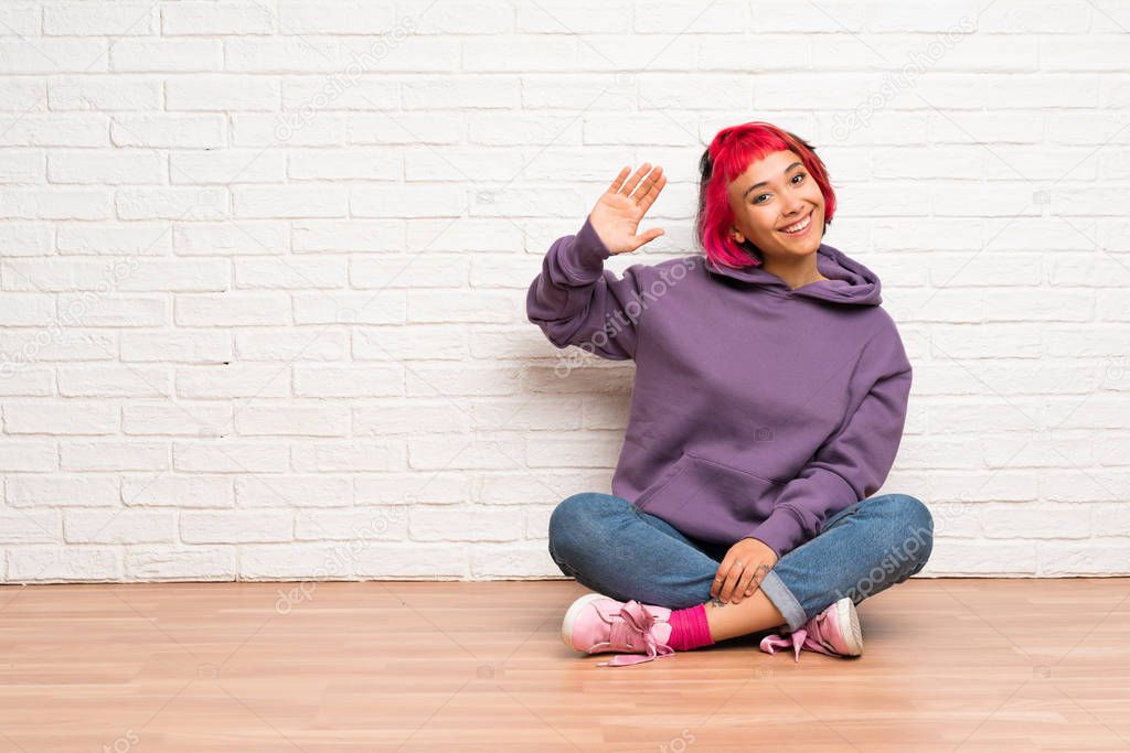 Young woman with pink hair sitting on the floor saluting with hand with happy expression
