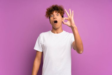 Genç Afro-Amerikan bir adam mor duvarın üstünde şaşırmış ve iyi bir işaret gösteriyor.