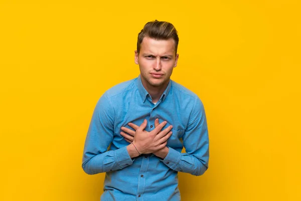 Blonde man over isolated yellow wall having a pain in the heart