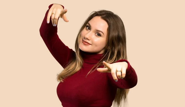 Teenager girl with turtleneck points finger at you while smiling over isolated ocher background
