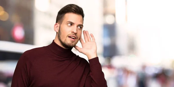 Man with turtleneck sweater listening to something by putting hand on the ear in the city