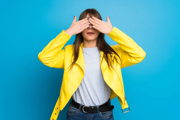 Young woman with yellow jacket on blue background covering eyes by hands. Surprised to see what is ahead