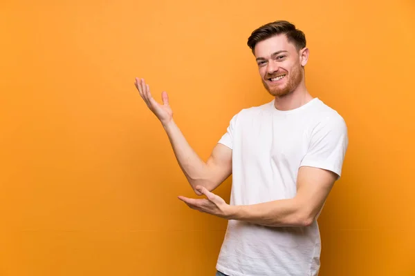 Redhead man over brown wall extending hands to the side for inviting to come