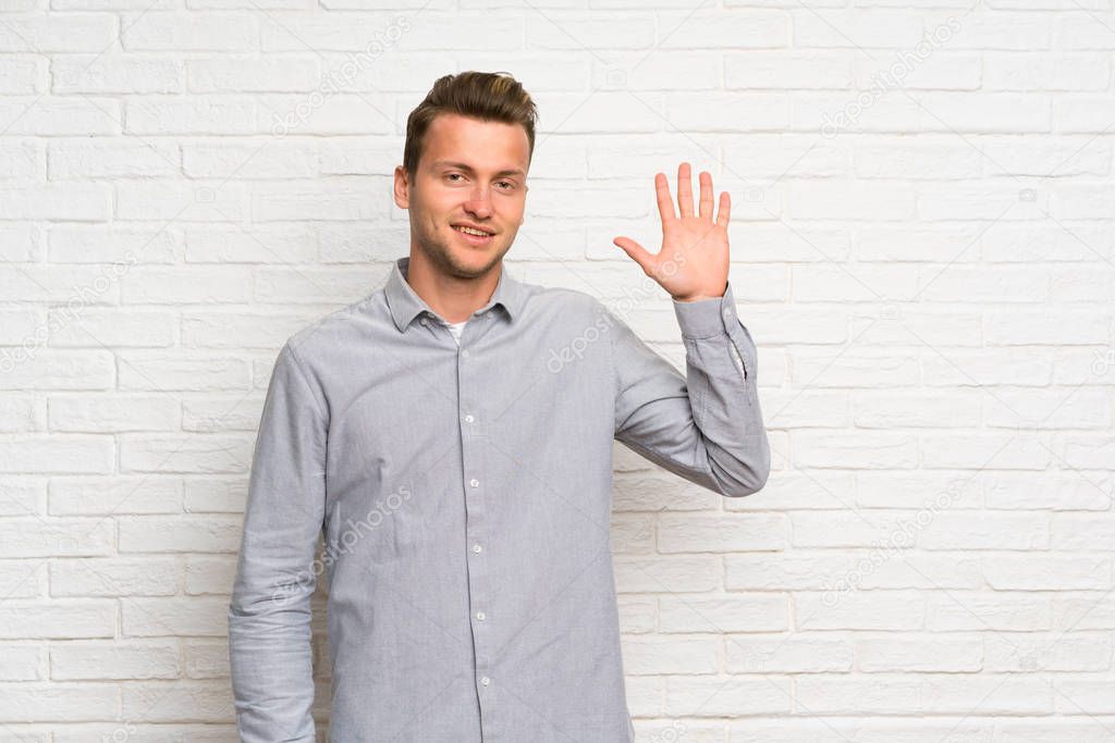 Blonde man over white brick wall saluting with hand with happy expression