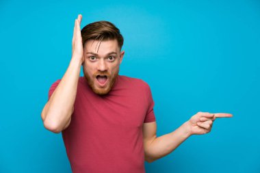 Izole mavi duvarda Redhead adam yan parmak işaret ve bir ürün sunan