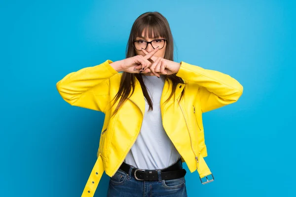 Young woman with yellow jacket on blue background showing a sign of silence gesture