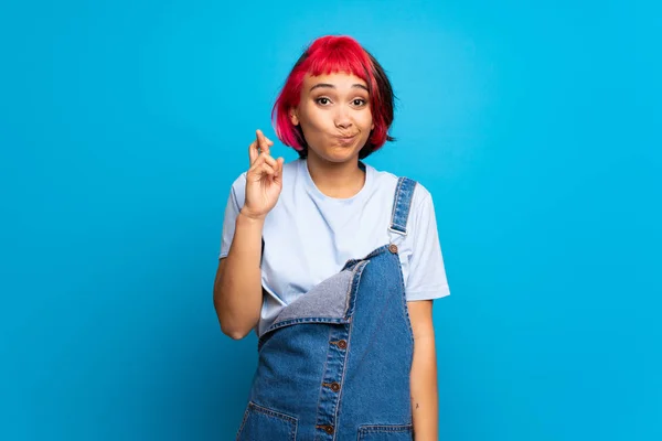 Young woman with pink hair over blue wall with fingers crossing and wishing the best