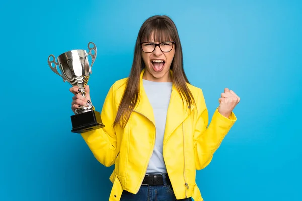 Young woman with yellow jacket on blue background holding a trophy