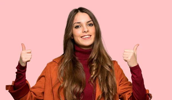 年轻的妇女与外套给竖起大拇指的姿态用双手和微笑在被隔绝的粉红色的背景 — 图库照片