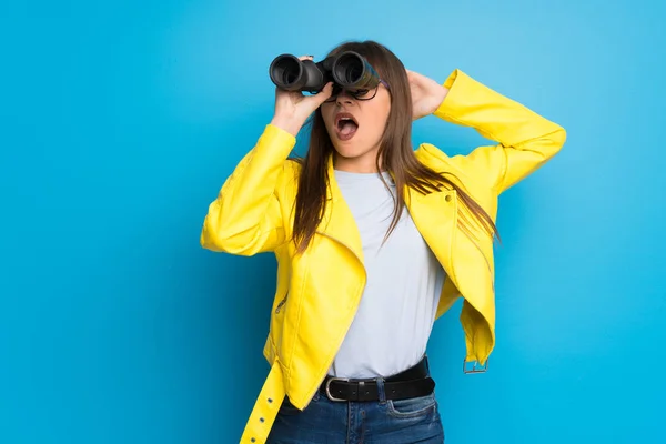 Young woman with yellow jacket on blue background and looking in the distance with binoculars