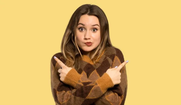 茶色のセーターが孤立した黄色の背景に疑問を持っている側面を指しているティーンエイジャーの女の子 — ストック写真