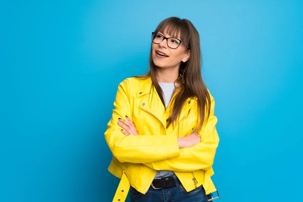 Young woman with yellow jacket on blue background keeping the arms crossed while smiling