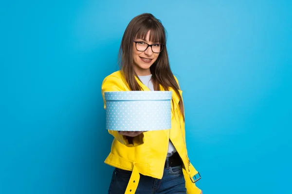 Young woman with yellow jacket on blue background holding a gift in hands