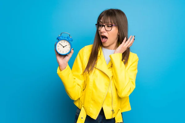 Jovem Mulher Com Casaco Amarelo Fundo Azul Segurando Relógio Alarme — Fotografia de Stock