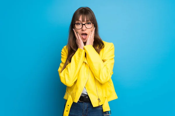 Young woman with yellow jacket on blue background surprised and shocked while looking right