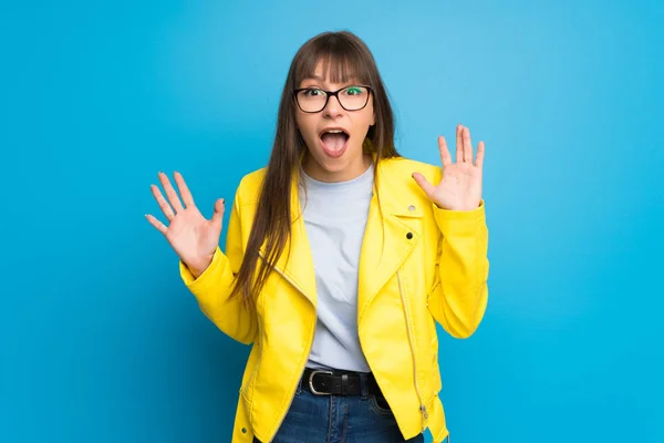 Young woman with yellow jacket on blue background with surprise and shocked facial expression