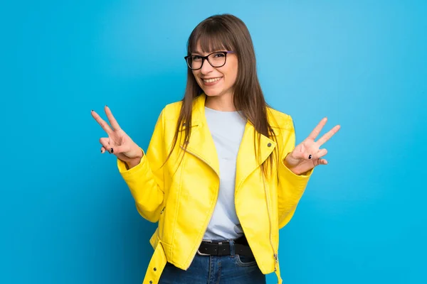 Young woman with yellow jacket on blue background smiling and showing victory sign with both hands