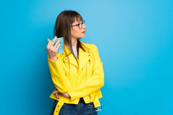 Young woman with yellow jacket on blue background holding a hot cup of coffee