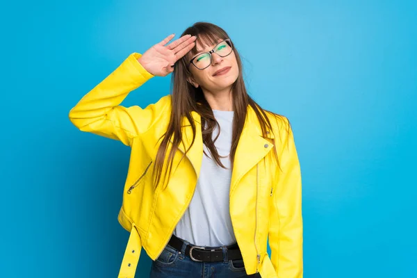 Young woman with yellow jacket on blue background saluting with hand