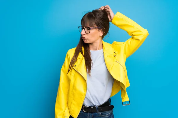 Young woman with yellow jacket on blue background having doubts while scratching head