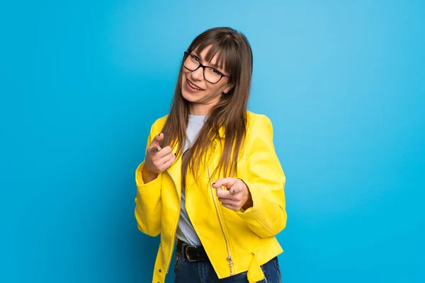 Young woman with yellow jacket on blue background pointing to the front and smiling