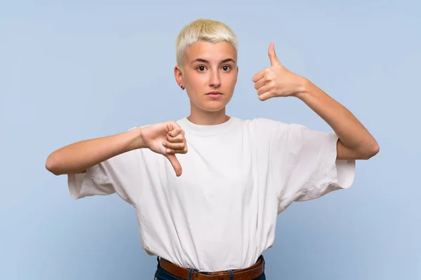Teenager girl with white short hair over blue wall making good-bad sign. Undecided between yes or not