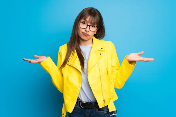 Young woman with yellow jacket on blue background having doubts while raising hands and shoulders