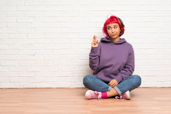 Young woman with pink hair sitting on the floor with fingers crossing and wishing the best