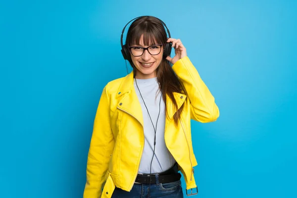 Young woman with yellow jacket on blue background listening to music with headphones
