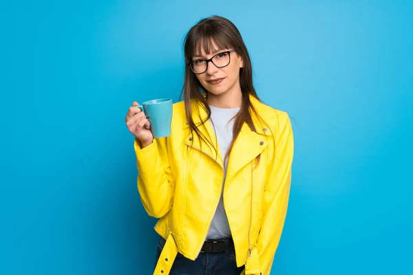 Young woman with yellow jacket on blue background holding a hot cup of coffee