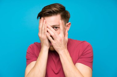 Gözleri kaplayan ve parmaklardan bakan izole mavi duvarda Redhead adam