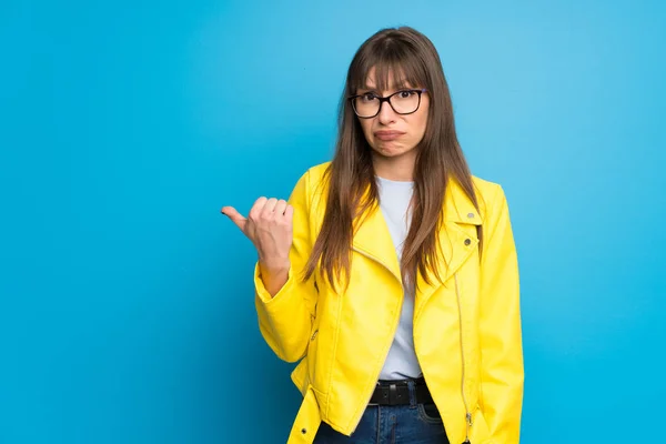 Young woman with yellow jacket on blue background unhappy and pointing to the side