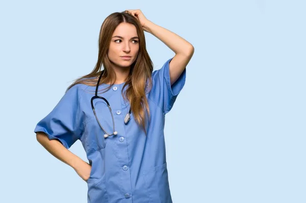在孤立的蓝色背景上挠头时感到疑惑的年轻护士 — 图库照片