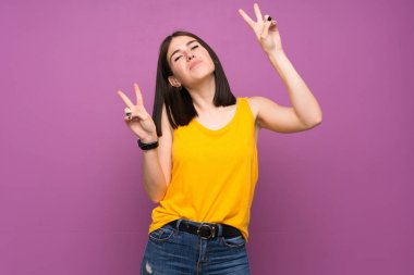 İzole edilmiş mor duvarlı genç bir kadın iki eliyle zafer işareti gösteriyor.