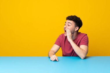 Renkli duvarı ve masası olan genç bir adam ağzı sonuna kadar açık bağırıyor.