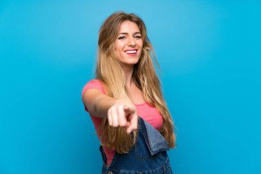 Önlüklü, mavi duvarlı genç sarışın kadın kendini beğenmiş bir ifadeyle seni işaret ediyor.