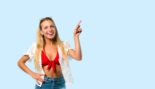 夏休みのブロンドの女の子は 側面に指を指し 青い背景に微笑みながら製品を提示 — ストック写真