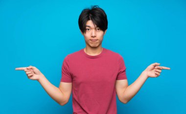 Izole mavi duvar üzerinde kırmızı gömlek li Asyalı adam şüpheleri olan lateraller işaret