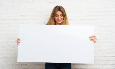 Beyaz tuğlalı duvarın üzerinden geçen genç sarışın kadın bir konsept eklemek için boş beyaz bir levha tutuyor.