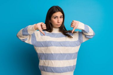 Mavi duvarın üzerinde iki eliyle başparmak aşağı gösteren genç kadın