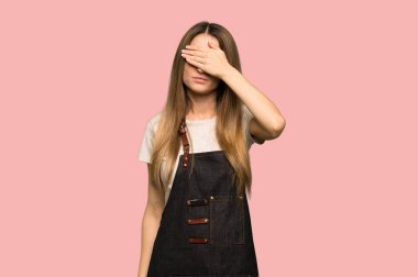Young woman with apron covering eyes by hands. Do not want to see something on isolated pink background