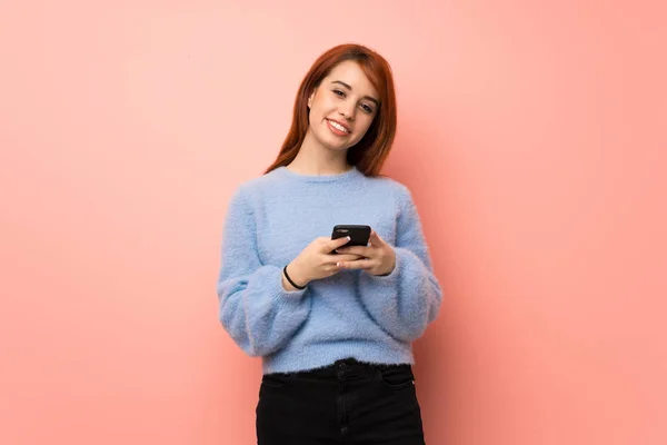 Young redhead woman over pink background sending a message with the mobile